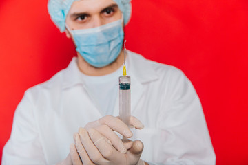 The doctor holds a syringe with a vaccine in his hands. A young guy in a white coat, in a medical mask and gloves on a red background. Medical worker.