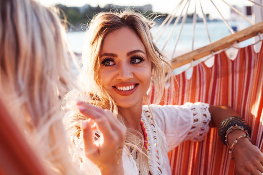 Two young happy beautiful girls dressed boho style white clothes, bracelets sit in hammock at the background sun and ocean