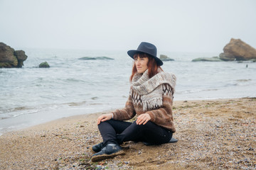 Beautiful woman in a hat and scarf sits on the coast against the background of the rocks against the beautiful sea. Place for text or advertising