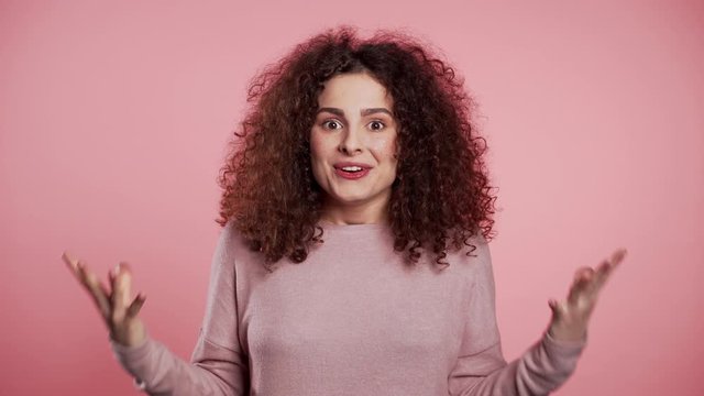 Portrait of girl with curly hair, she shows wow delight effect gesture. Surprised excited happy woman. Pretty female shocked model on pink background.