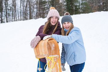 Two happy teen girls/friends standing behind a wood sled/toboggan while standing at the bottom of hill covered with snow.