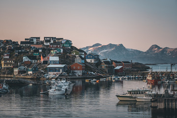 Fototapeta na wymiar Sunset view of Maniitsoq arctic city in Greenland. Mountains in background during midnight sun. Colourfull houses and panorama. View to port with ships.