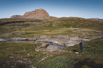 Fototapeta na wymiar Young man hiker in the arctic landscape of Disko Bay in Greeland in Summer. Blue Sky and green meadows. Arctic Circle Trail with temple mountains. Disko Island and Village of Qeqertarsuaq,