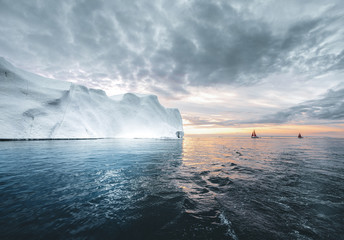 Beautiful red sailboat in the arctic next to a massive iceberg showing the scale. Cruising among...