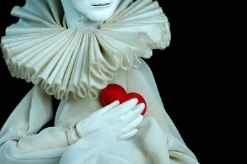 .Harlequin doll holds a small red heart for Valentine's Day on a black background. - obrazy, fototapety, plakaty