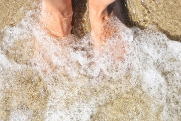 Fototapeta premium Woman’s bare feet at the transparent sea water with golden sand, selective focus. Bare feet in a sea wave. Relaxation in the ocean. Woman feet under the water. Summertime. 