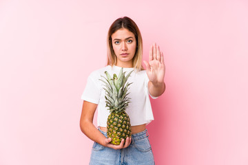 Young caucasian woman holding a pineapple standing with outstretched hand showing stop sign,...