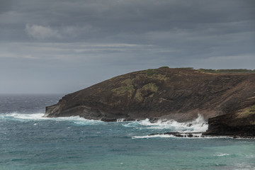 Fototapeta premium Oahu, Hawaii, USA. - January 11, 2020: Hanauma Bay Nature Preserve. Baboon face cliff sits at entrance of bay with greenish water. Darker ocean water behind and very heavy dark cloudscape.