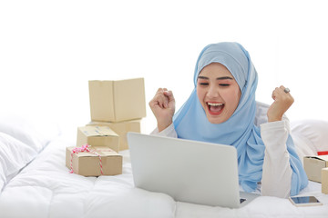 Beautiful and young asian muslim woman in sleepwear with attractive look, lies on bed with computer, mobile phone and online package box delivery. Smart girl with hijab receive good news and surprise.