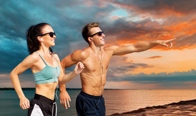 fitness, sport and technology concept - happy couple with earphones running over sea and sunset sky background