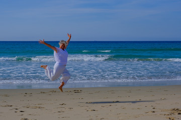 Senior woman with greay hair and a white dress jumping in the air at the beach