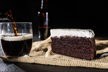 Portion of chocolate cake and black beer on black background with bottle and serving black beer in glass - obrazy, fototapety, plakaty