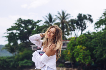 Naklejka premium Tanned caucasian blonde young woman in a white shirt is enjoying the sun on the background of a palm trees. Paradise on earth