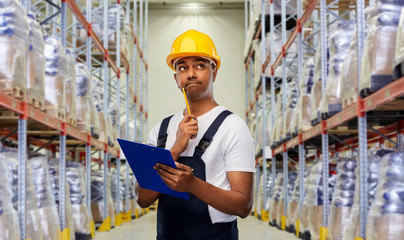 logistic business and people concept - happy smiling indian loader or worker in helmet with clipboard and pencil over warehouse background