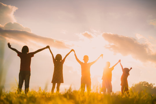 The Silhouette Of The Children Holding Hands, Enjoying The Sunset, A Group Of Friends Cheering And Arms Raised In The Orange Sky And The Mountains Behind Friendship Concept