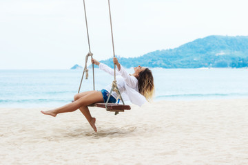 Tanned beautiful caucasian blonde young woman swinging on a wooden swing on the shore of the blue sea. Sunny day, white sand. Paradise on earth. White shirt fluttering