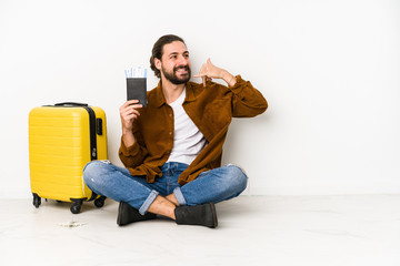 Young caucasian man sitting holding a passport and a suitcase isolated showing a mobile phone call gesture with fingers.