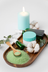 Fototapeta na wymiar beauty and spa concept - green bath salt, serum with dropper or essential oil, moisturizer and eucalyptus cinerea with cotton flowers on wooden tray