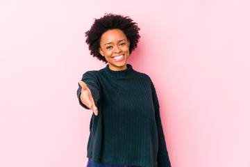 Middle aged african american woman against a pink background isolated stretching hand at camera in greeting gesture.