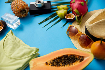 summer holidays, vacation and travel concept - close up of sunglasses, straw hat, camera and exotic fruits with palm leaf and clothes on blue background