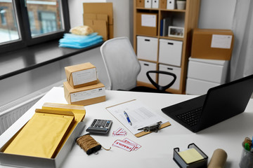 Naklejka premium delivery, mail service and shipment concept - laptop, clipboard, parcel boxes and packing stuff on table at post office