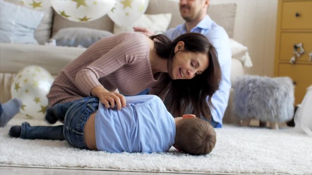 Happy mom is tickling her little laughing son on the floor at living room, close-up. Father playing with children on the background