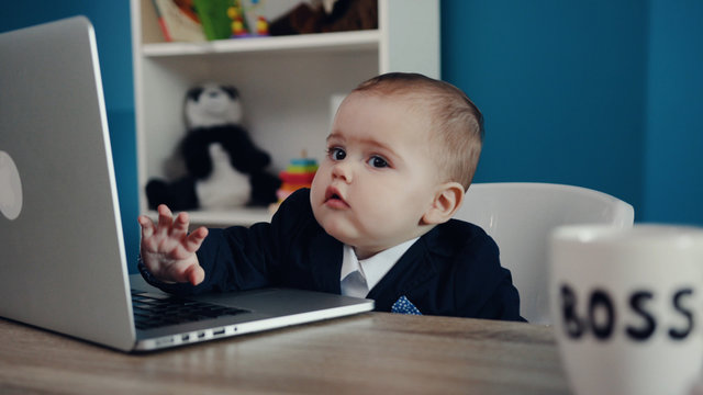 Photo of adorable serious baby boss in business suit typing on keyboard of the laptop while sitting in the modern office. Success, happy childhood, cuteness. Child&rsquo;s portrait. Close up view