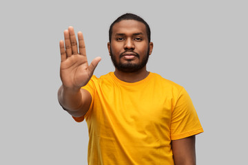 warning and people concept - young african american man in yellow t-shirt showing stop gesture over grey background