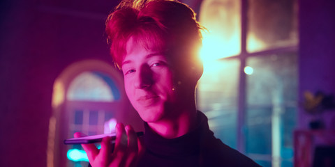 Recording voice message. Cinematic portrait of redhair man in neon lighted interior. Toned like cinema effects in purple-blue. Caucasian model using smartphone in colorful lights indoors. Flyer.