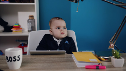 Lovely toddler boy in business suit with extremely cute cheeks sits at the table with tablet by the baby boss mug. Successful baby, childhood happiness, little wonder.