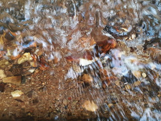Ripples in the river flow through the rocks, colorful in the summer in bright sunlight, have background of the shiny pebbles, at the bottom of river In a abstract, form of pebbles below water surface.