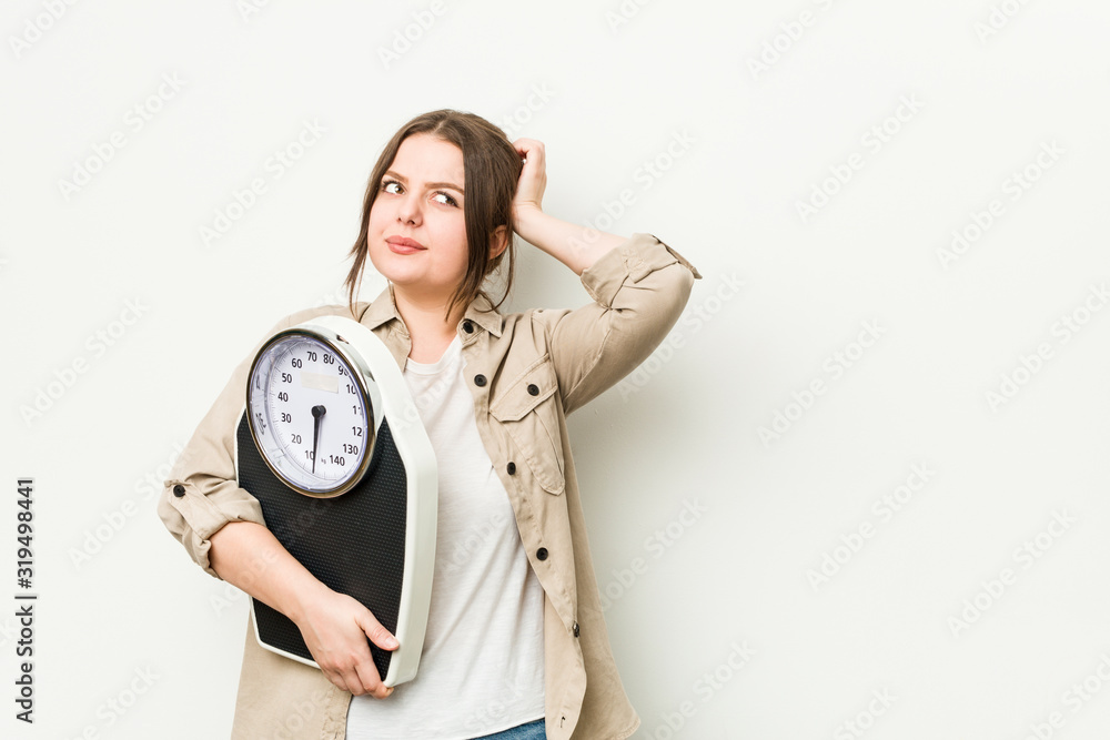 Wall mural young curvy woman holding a scale touching back of head, thinking and making a choice.