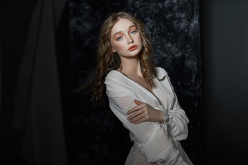 Romantic portrait of a young blonde woman in a light white dress. Girl with perfect skin poses on a dark background
