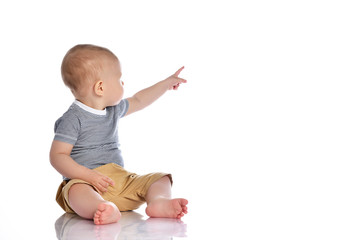 Infant baby boy in t-shirt and pants is sitting on the floor looking back and pointing at upper corner behind him