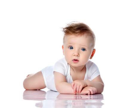 Surprised intrigued infant child baby boy toddler with blue eyes in white bodysuit lies on the floor on his stomach