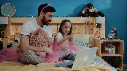 Fairytale family party at home. View of happy father and daughter playing fairies waving with magic wand looking on laptop screen sitting on bed and having fun.