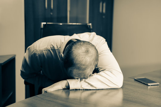 Middle aged man is depressed and exhausted. He just sitting at working place with his head on the table. Sepia color.