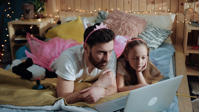 Side view of happy father and daughter dressed like fairies watching cartoons on laptop. Nice funny young family spending time together at magic party at home.