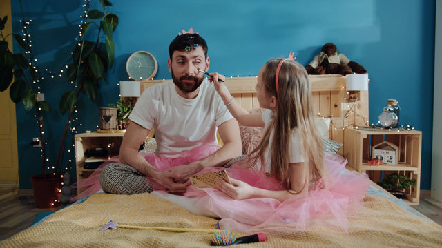 Family preparation for birthday fancy party. Little girl fairy applying makeup to her father. Portrait of silly happy young man smiling and sitting on bed. Two pretty magic fairies.