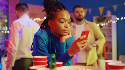 Unsociable afro-american glamorous girl browsing news on smartphone staying online while her friends dancing and having fun. Outsider. Concept of nomophobia.