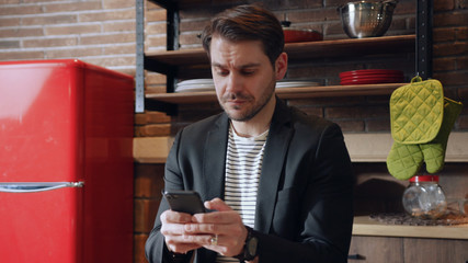 Attractive young man in formal suit focused on his smartphone and messaging app, actively typing the sms while sitting at table in the kitchen. Social networks, internet usage, texting. Addiction and