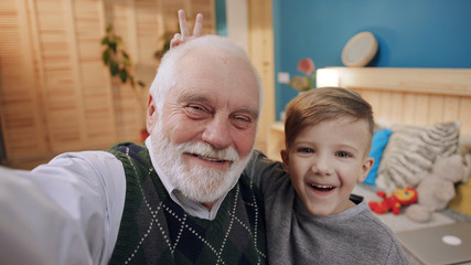 Happy grandfather and grandson taking a selfie together sitting on sofa waving with a hand smiling...