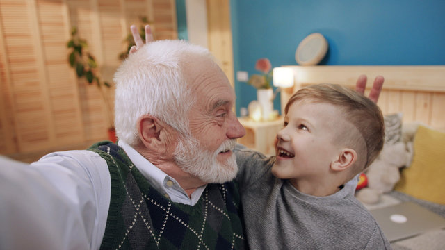 Funny grandfather and grandson taking a selfie together sitting on sofa waving with a hand smiling family child grandchild grandparent old indoors senior childhood fun senior close up slow motion