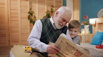 Nice child holding on to hand of grandpa and men reading newspaper together. Close-up footage from below. Indoors. Bedroom. Lifestyle. Love, daily, concept.