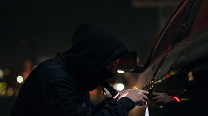 Close up robber checking breaking entering alarm shines a flashlight in a car sitting stealing at...