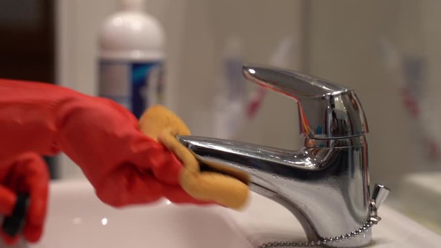 Housewife in protective rubber orange gloves cleaning stainless water tap with yellow sponge and detergent gel. Dirty bathroom faucet, household chemicals, hygiene, sanitation and house cleaning