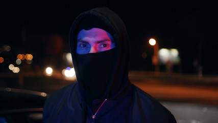 Man bandit in balaclava and hood looking at the camera at night blinking headlights colorful danger...