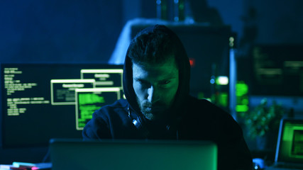 Portrait of concentrated dangerous hacker coding virus ransomware on laptop and multiple displays...