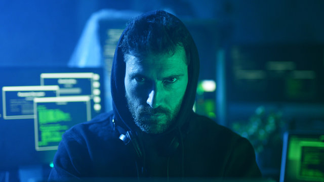 Portrait of menacing serious computer hacker working with codes on laptop at desk. Cyber crime and malware concept.