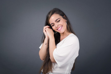 Dreamy young female with pleasant expression, closes eyes, keeps hands crossed near face, thinks about something pleasant, poses against gray background.